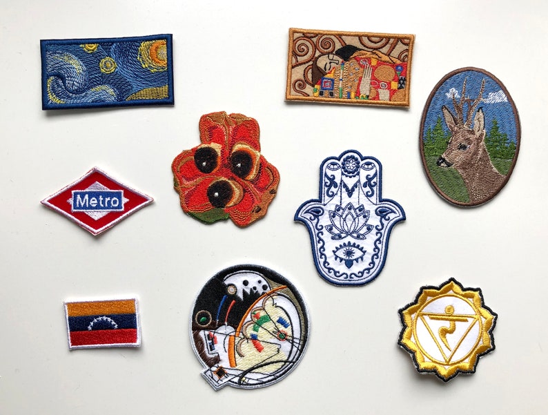 May include: A collection of embroidered patches with various designs. Included are patches depicting a starry night scene, a deer in an oval frame, a red and white Metro sign, a red flower, a Hamsa hand, a flag, a circular art piece, and a yellow chakra symbol.