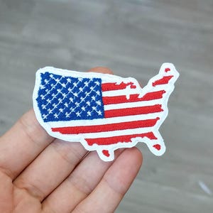 May include: An embroidered fabric patch shaped like the contiguous United States. The design features the American flag pattern with a blue field containing white stars on the left and horizontal red and white stripes across the rest of the map.