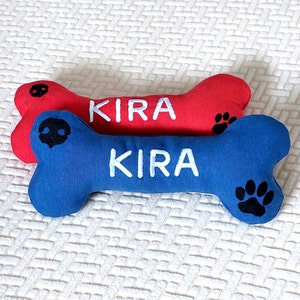 Könnte beinhalten: Zwei Plüsch-Hundespielzeuge in Knochenform. Der obere Knochen ist rot mit dem Namen "KIRA" in weiß gestickt. Der untere Knochen ist blau mit dem gleichen Namen in weiß gestickt. Beide Knochen haben einen schwarzen Pfotenabdruck auf einer Seite.