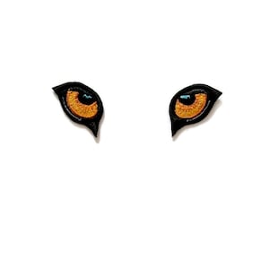 K&ouml;nnte beinhalten: Zwei schwarze gestickte Augen mit orangefarbenen Iris und blauen Pupillen. Die Augen befinden sich auf einem wei&szlig;en Hintergrund.