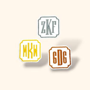 Könnte beinhalten: Drei gestickte Monogramm-Patches in quadratischer Form mit dekorativem Rand. Die Patches zeigen die Initialen ZKF in Grau, MKW in Gelb und GDG in Braun. Jeder Patch befindet sich auf einem cremefarbenen Hintergrund.