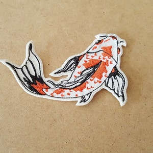 May include: Embroidered patch featuring a koi fish design. The fish has a white body with orange spots and black outlines. The tail and fins are detailed with black and white patterns. The patch is likely for clothing or accessories.