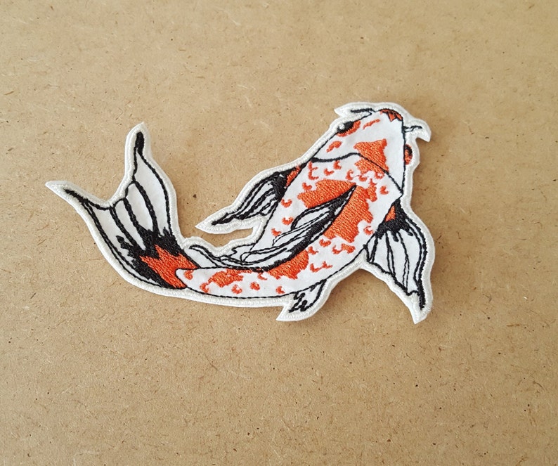 May include: Embroidered patch featuring a koi fish design. The fish has a white body with orange spots and black outlines. The tail and fins are detailed with black and white patterns. The patch is likely for clothing or accessories.