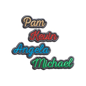 Puede incluir: Cuatro parches bordados para planchar con los nombres "Pam", "Kevin", "Angela" y "Michael" en diferentes colores. Los parches están sobre un fondo blanco.