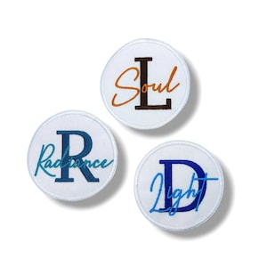 Puede incluir: Tres parches bordados circulares blancos. Un parche tiene la palabra "Soul" con una "L" marrón. Otro parche tiene la palabra "Radiance" con una "R" azul. El tercer parche tiene la palabra "Light" con una "D" azul.