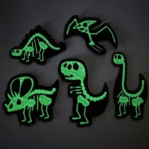 May include: Five black dinosaur patches with glow-in-the-dark green skeleton designs. The patches feature a Stegosaurus, Pterodactyl, Triceratops, T-Rex, and a Brontosaurus. These patches are ideal for adding a unique touch to clothing or accessories.
