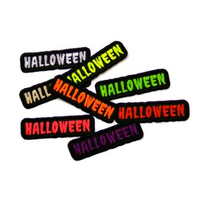 Op de afbeelding: Een verzameling van acht zwarte viltpatches met het woord "Halloween" geborduurd in verschillende kleuren, waaronder wit, groen, oranje, rood en paars.