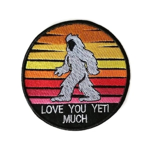 以下が含まれることがあります： 白いイエティが夕焼けの前を歩いている、白い文字で「Love You Yeti Much」と書かれた丸い刺繍パッチです