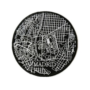 Può includere: Toppa ricamata in bianco e nero di una mappa di Madrid, Spagna. La toppa è circolare e ha un contorno bianco delle strade della città e la parola "MADRID" in lettere bianche.