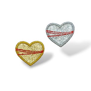 Peut inclure: Deux écussons en forme de cœur. L'un est doré avec une bande horizontale rouge, et l'autre est argenté avec une bande horizontale rouge. Les deux ont une texture pailletée et sont posés sur un fond blanc.