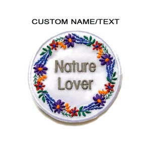Peut inclure: Patch brodé blanc avec un motif de couronne de fleurs. La couronne est composée de fleurs violettes, orange et vertes. Le texte "Nature Lover" est au centre du patch.