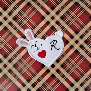 May include: A white heart-shaped patch featuring a bunny face, a red heart, and the letter "R". The patch has a white background with pink ears and a fluffy tail. The background is a plaid pattern in red, black, and tan.