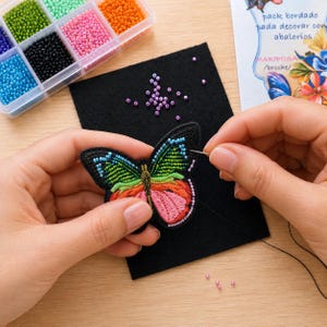 May include: A colorful beaded butterfly applique with blue, green, and pink wings is being sewn onto black fabric. A clear plastic organizer filled with various colored beads sits in the background, along with a card that says "pack bordado para decorar con abalorios".