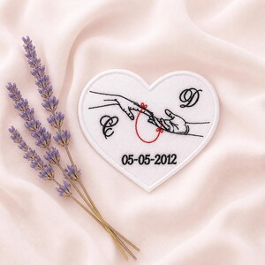 May include: A white heart-shaped patch with black embroidered hands connected by a red string. The patch includes the initials "C" and "D" and the date "05-05-2012". Lavender sprigs are arranged on a pink fabric background.
