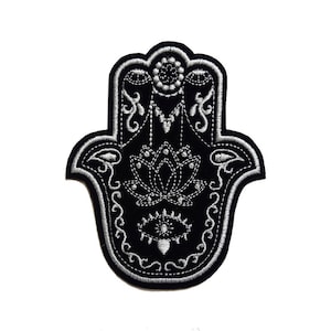 May include: Black and white embroidered patch of a hamsa hand with a lotus flower and an eye in the centre.