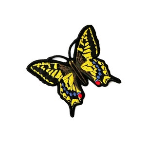 Puede incluir: Un parche bordado de mariposa amarillo y negro con detalles azules y rojos. La mariposa está en vuelo con las alas extendidas.