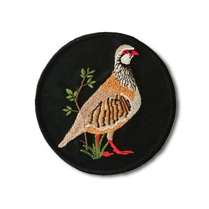 Peut inclure: Patch brodé représentant une perdrix brune et blanche debout sur un buisson vert sur fond noir.