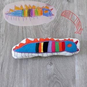 May include: A colourful, embroidered caterpillar cushion with a blue body, orange and red spikes, and a variety of coloured stripes. The cushion is made of white fabric and is lying on a wooden surface.