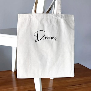 May include: Cream-colored tote bag with the word "Dreams" in black script. The bag has a simple design with two handles and is made of a canvas-like material. The bag is hanging on a white chair.