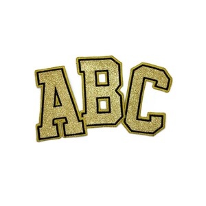 Op de afbeelding: Gouden glitterletters die "ABC" spellen met zwarte contouren. De letters zijn in een varsity-stijl lettertype.
