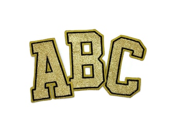 以下が含まれることがあります： 黒いアウトラインで「ABC」と綴られた金色のグリッターレター。レターはバーシティスタイルのフォントです。