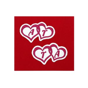 May include: Two pink intertwined hearts with white outlines and the letters "AT" and "TA" embroidered inside.