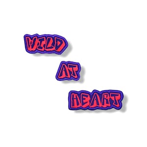 Peut inclure: Trois écussons brodés avec les mots "WILD", "AT" et "HEART" dans une police de style graffiti. Le texte est rose vif avec un contour violet. Les écussons sont disposés en diagonale sur un fond blanc.