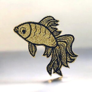 May include: A glittery gold and black goldfish design. The fish has a detailed outline in black, with gold glitter filling the body. The fins and tail are also black, creating a striking contrast. The overall design is whimsical and decorative.