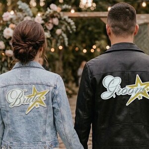 Pode incluir: Um casal em um casamento, visto por trás, de mãos dadas. A pessoa à esquerda usa uma jaqueta jeans azul clara com um patch "Bride". A pessoa à direita usa uma jaqueta de couro preta com um patch "Grooms". Os patches apresentam um design de estrela.