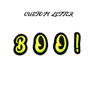 Può includere: Un set di quattro toppe da stirare che compongono la scritta "BOO!" in uno schema di colori giallo e nero. Le lettere sono in un font stilizzato.