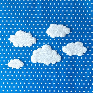 Può includere: Cinque decorazioni a forma di nuvola bianca e soffice con una superficie strutturata sono disposte su uno sfondo blu a pois bianchi. Le nuvole variano di dimensioni, creando un'estetica stravagante e giocosa. L'immagine è luminosa e allegra.