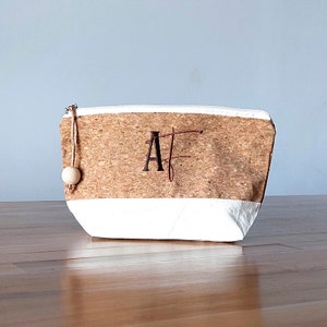 May include: A cork and canvas zippered pouch with a white bottom and a brown cork top. The pouch has a wooden bead zipper pull and the initials "AF" embroidered on the cork.