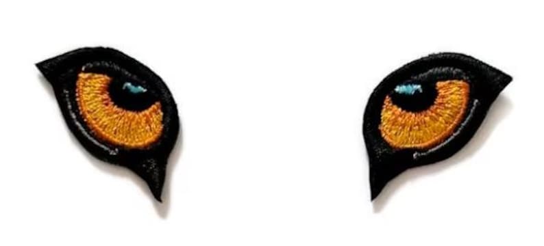 K&ouml;nnte beinhalten: Zwei gestickte B&uuml;gelbilder mit einem schwarzen Katzenauge mit orangefarbener Iris und blauer Pupille.