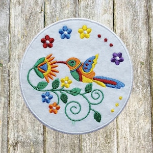 May include: Embroidered white circular patch featuring a colorful hummingbird with a long beak, drinking nectar from a flower. The hummingbird has blue, yellow, red, and green details. Surrounding the hummingbird are embroidered flowers in various colors.