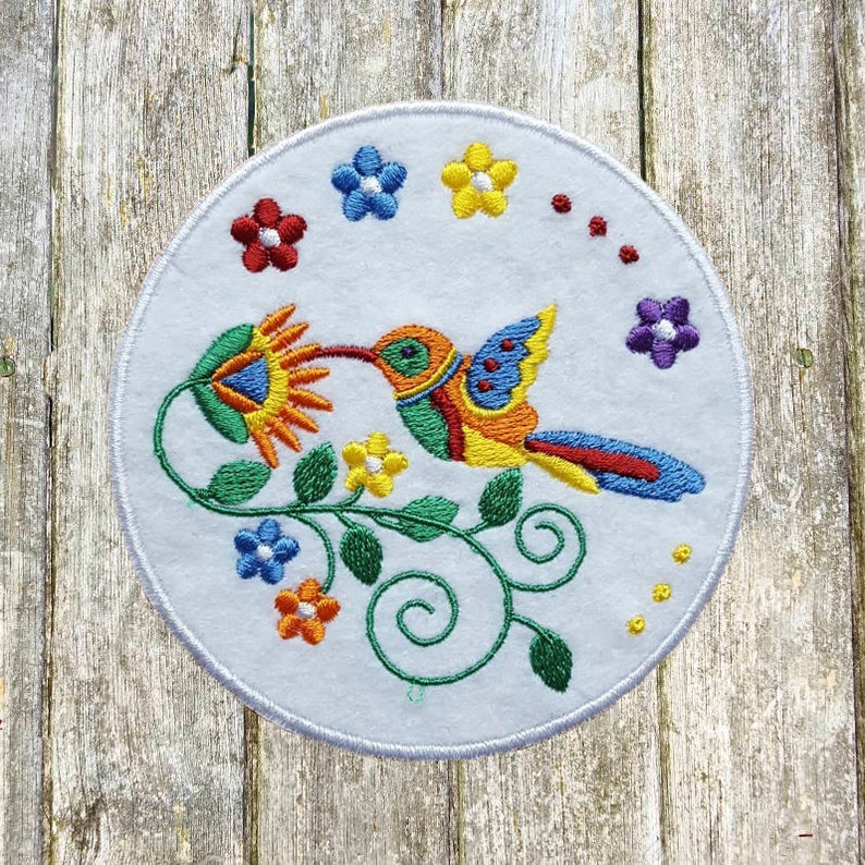 May include: Embroidered white circular patch featuring a colorful hummingbird with a long beak, drinking nectar from a flower. The hummingbird has blue, yellow, red, and green details. Surrounding the hummingbird are embroidered flowers in various colors.