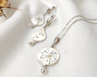 Puede incluir: Un conjunto de collar y pendientes de plata con un delicado diseño de diente de león. El colgante y los pendientes presentan un diente de león de esmalte blanco con detalles plateados.