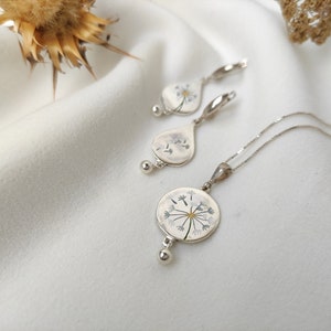 May include: A silver necklace and earrings set with a delicate dandelion design. The pendant and earrings feature a pressed dandelion flower with white petals and a yellow center.