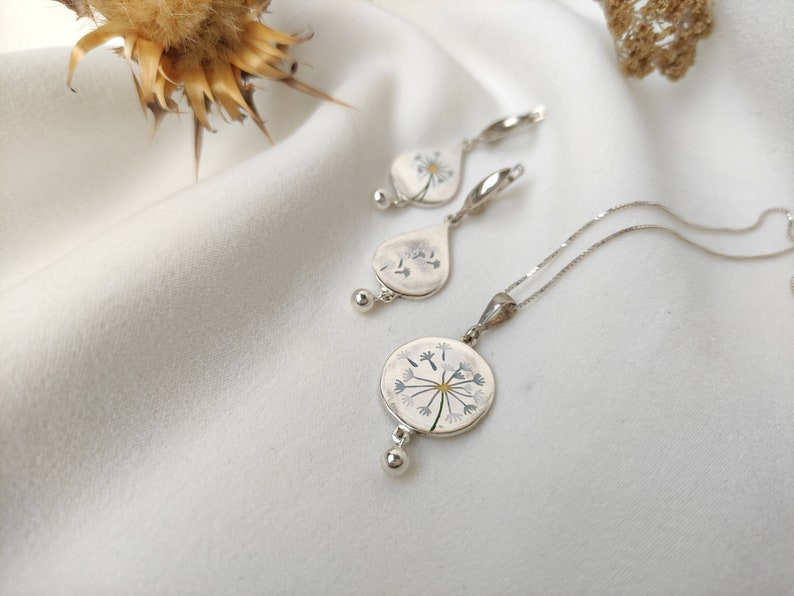 May include: A silver necklace and earrings set with a delicate dandelion design. The pendant and earrings feature a pressed dandelion flower with white petals and a yellow center.