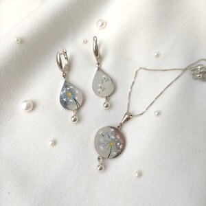 May include: A silver necklace and earrings set with a dandelion design. The pendant and earrings feature a white dandelion with a yellow center.
