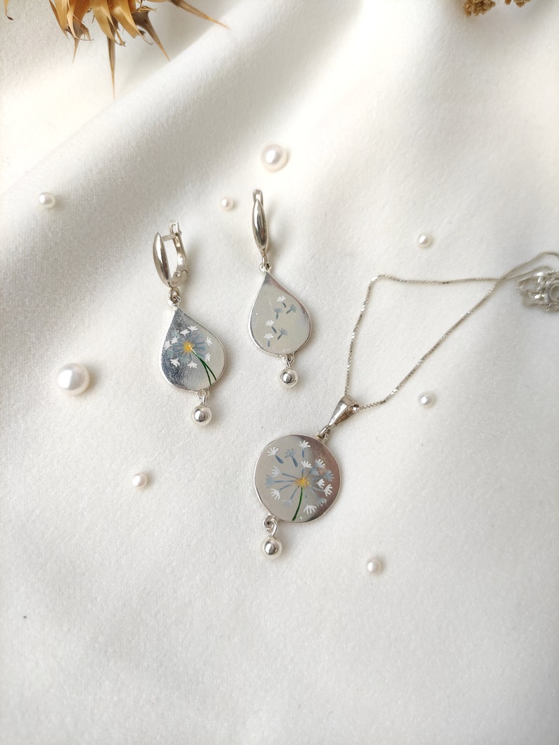 May include: A silver necklace and earrings set with a dandelion design. The pendant and earrings feature a white dandelion with a yellow center.
