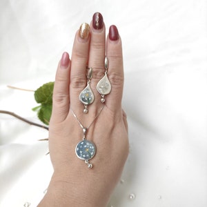 May include: A silver necklace and earrings set with a delicate flower design. The pendant is a round circle with a flower pressed inside. The earrings are teardrop shaped with a flower pressed inside.