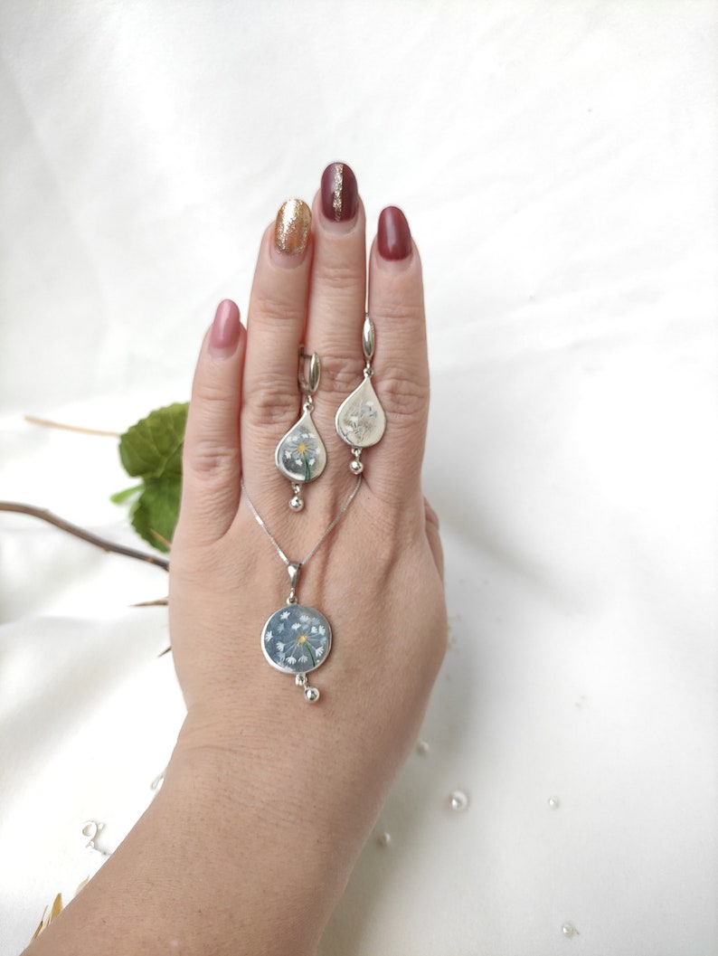 May include: A silver necklace and earrings set with a delicate flower design. The pendant is a round circle with a flower pressed inside. The earrings are teardrop shaped with a flower pressed inside.