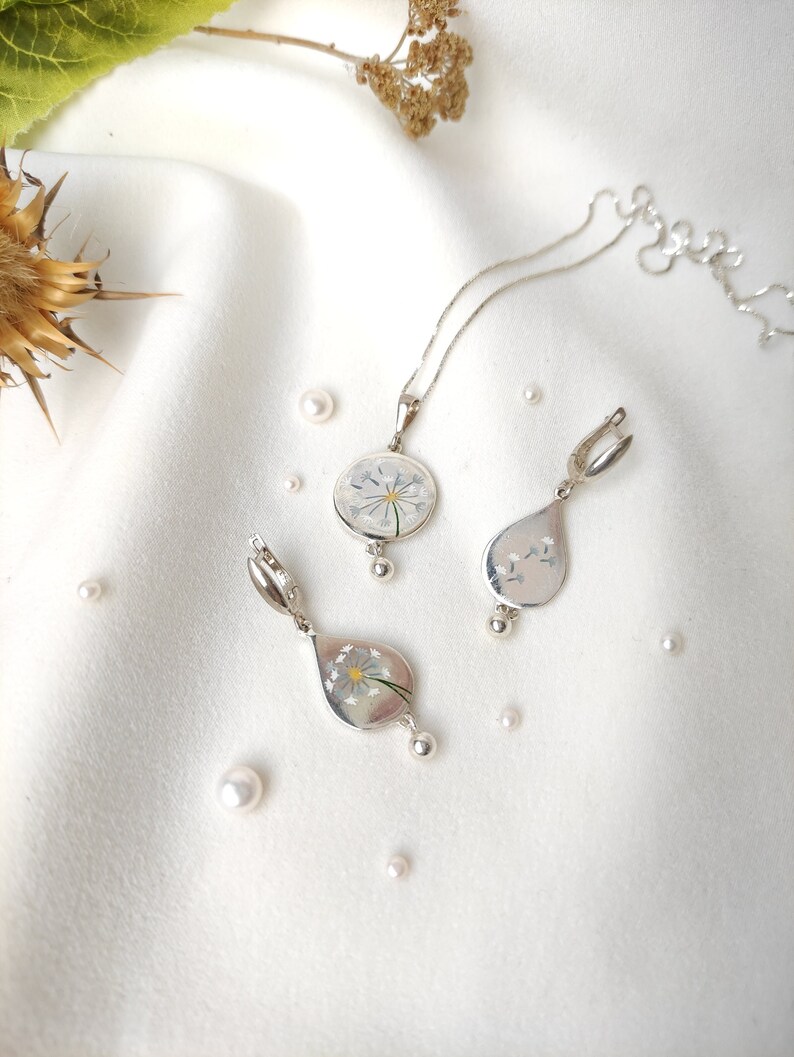 May include: A silver necklace and earrings set with a delicate flower design encased in clear resin. The pendant is round and the earrings are teardrop shaped.