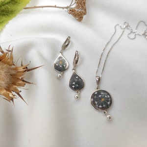 May include: A silver necklace and earrings set with a delicate dandelion design. The pendant and earrings feature a clear resin with a pressed dandelion flower.