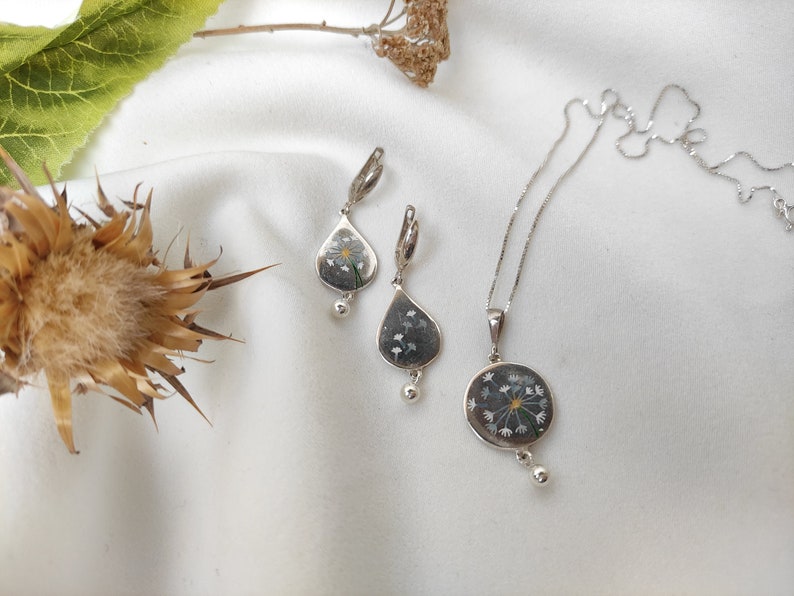 May include: A silver necklace and earrings set with a delicate dandelion design. The pendant and earrings feature a clear resin with a pressed dandelion flower.