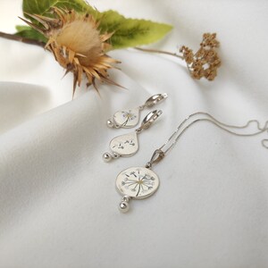 May include: A silver necklace and earrings set featuring a delicate dandelion design with white flower petals and a gold center. The jewelry is displayed on a white fabric background.