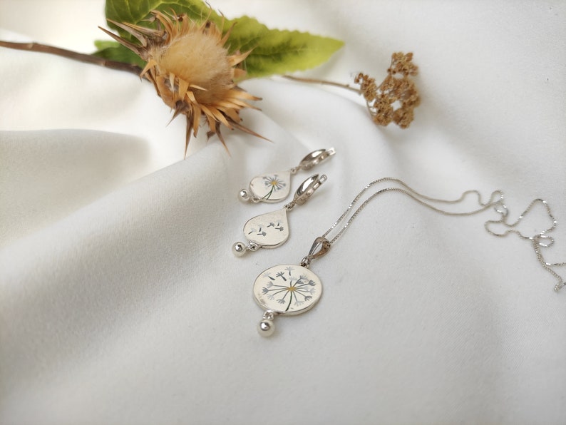 May include: A silver necklace and earrings set featuring a delicate dandelion design with white flower petals and a gold center. The jewelry is displayed on a white fabric background.