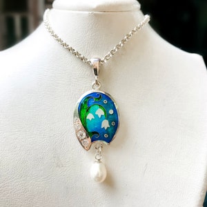 May include: A silver necklace with a pendant featuring a blue and green enamel design of lily of the valley flowers and a single pearl drop.