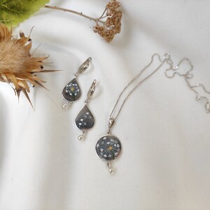 May include: A silver necklace and earrings set with a delicate flower pendant and matching teardrop earrings. The flower pendant is made with real pressed flowers and has a silver chain.