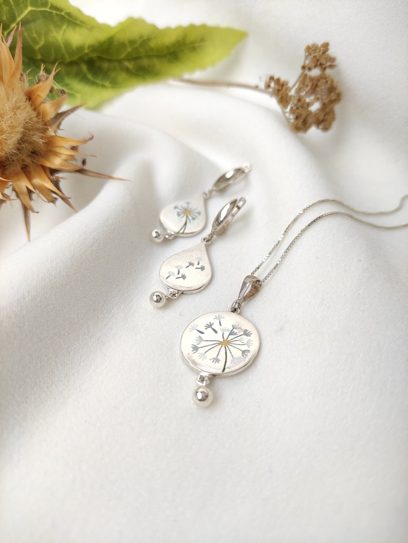 May include: A silver necklace and earrings set with a delicate dandelion design. The pendant and earrings feature a white enamel dandelion with silver details.